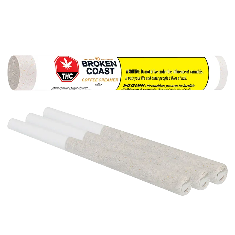 Angled close-up of three rolled joints below a horizontal Broken Coast Coffee Creamer PR 3 x 0.5 g tube with bold yellow warning label.