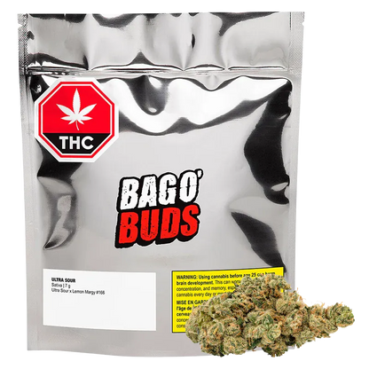 Front-facing shot of a silver pouch and bud labeled BAGO' BUDS Ultra Sour, with a red THC symbol at top left and a cannabis nugget resting at bottom right