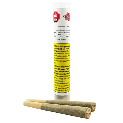 Tall front view of the Organic Apex Kush PR 2 x 1 g clear plastic tube with a yellow warning label, shown standing behind three pre-rolled joints.