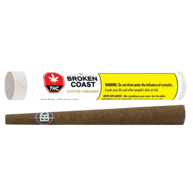 Horizontal product shot of a brown pre-rolled blunt below its white tube packaging labeled "Broken Coast Coffee Creamer Blunt PR 1 x 1 g" with a yellow warning panel.