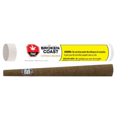 Horizontal product shot of a brown pre-rolled blunt below its white tube packaging labeled "Broken Coast Coffee Creamer Blunt PR 1 x 1 g" with a yellow warning panel.
