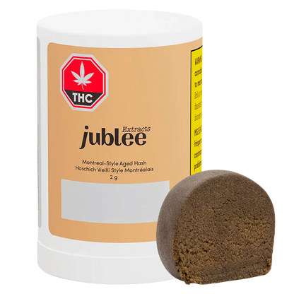 Front-facing shot of the Jubilee Extracts Montreal-Style Aged Hash 2 g cylinder with a brown half-disc of hash leaning against it.