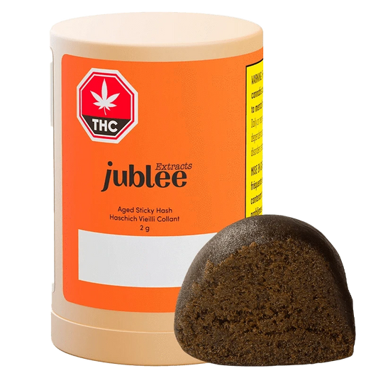 A close-up of the Aged Sticky Hash, showing a dark, dense block with a textured surface against a white background.