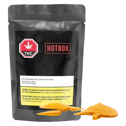 Front-facing shot of the black pouch labeled Hotbox House Shatter with a red THC badge and amber shatter pieces arranged at its base.