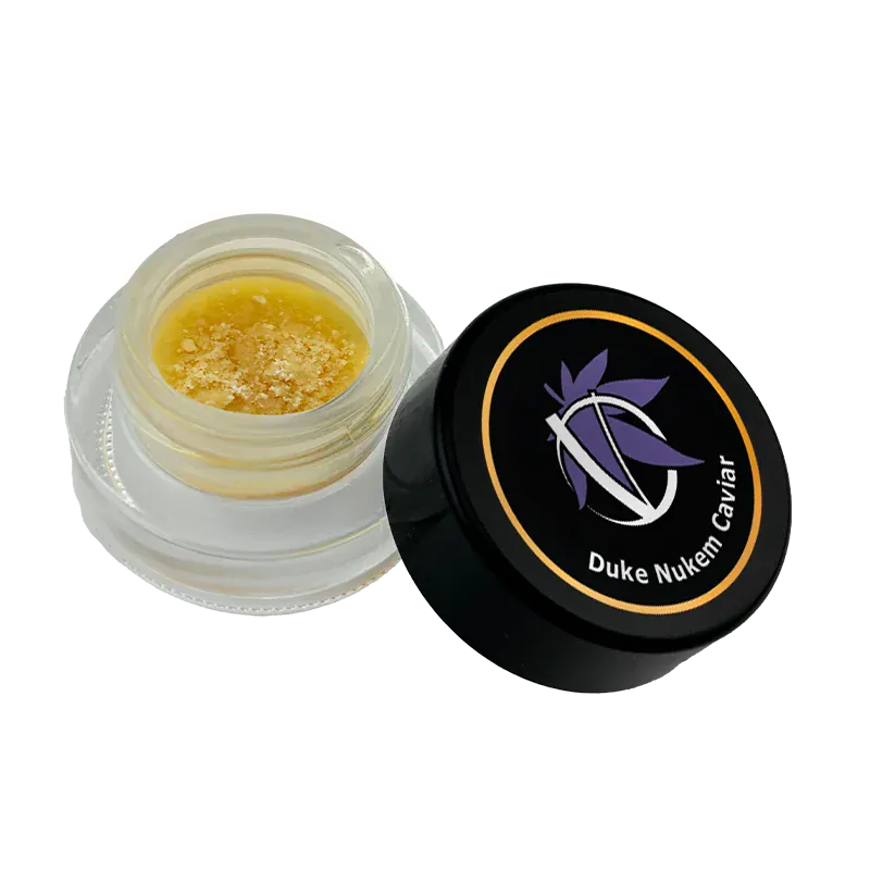 Angled top-down view of an open jar of Duke Nukem Caviar showing golden, crumbly concentrate inside and the black branded lid beside it.