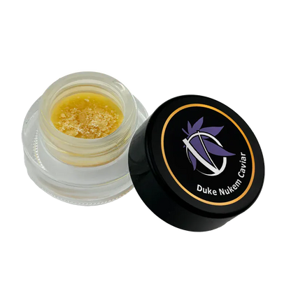 Angled top-down view of an open jar of Duke Nukem Caviar showing golden, crumbly concentrate inside and the black branded lid beside it.
