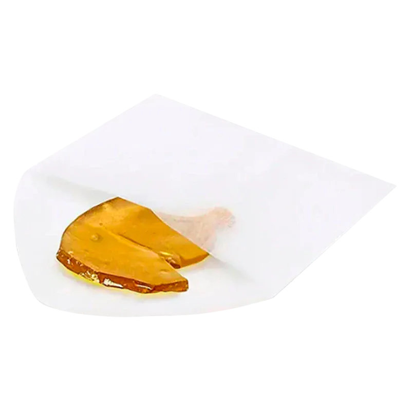 Close-up of a golden amber shard of Exhaust Haze Shatter partially wrapped in translucent white parchment paper on a white background.