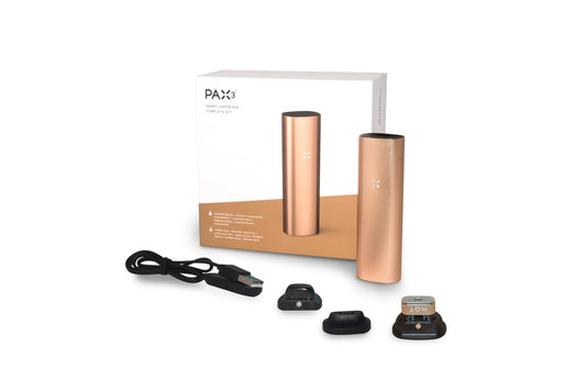 A close-up of the rose gold Pax 3 - Complete Kit, angled slightly, shows its sleek, metallic body and included accessories on a dark background.