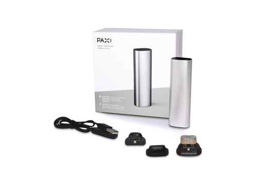 A studio shot displays the silver Pax 3 - Complete Kit, including the device, charging cable, and replacement oven lids on a white surface.