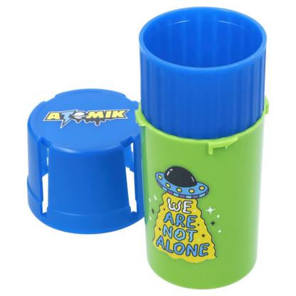 Close-up front view of the green and blue Atomik Grind & Stash tube with its blue lid beside it and a cartoon UFO “We Are Not Alone” graphic.