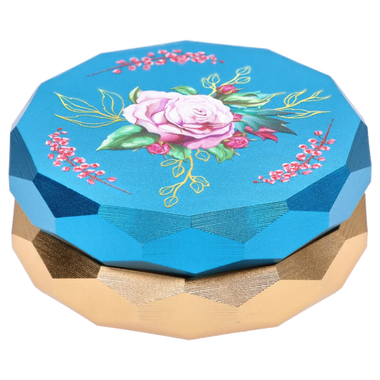 Top-down view of the Famous Design Floral Lush Aluminum 2 Stage Grinder - Teal and Gold showing a teal faceted lid with a central pink rose and gold-accented leaves above a metallic gold faceted base