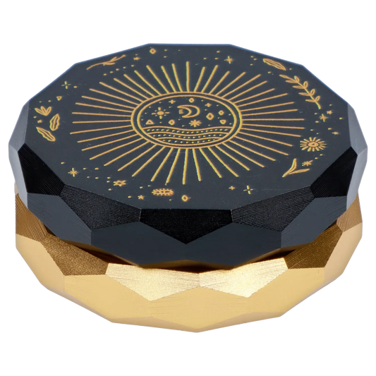 Top-down close-up of the Famous Design Astral Power Aluminum 2 stage Grinder -Black and Gold showing a faceted black lid with gold celestial sun and moon engraving atop a gold faceted base.