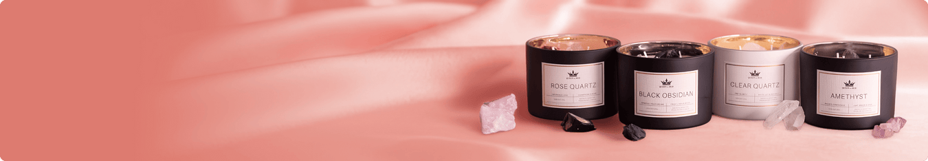 Four QOB crystal-themed candles (Rose Quartz, Black Obsidian, Clear Quartz, Amethyst) in black and white jars on pink satin with matching crystals