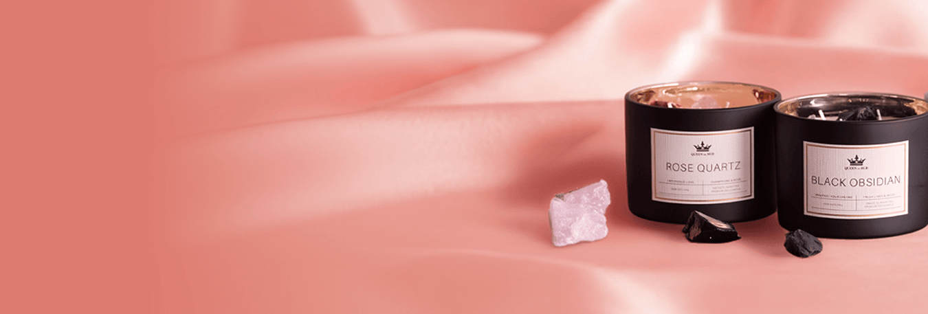 Two black QOB candles labeled "Rose Quartz" and "Black Obsidian" beside matching crystal stones on pink satin background