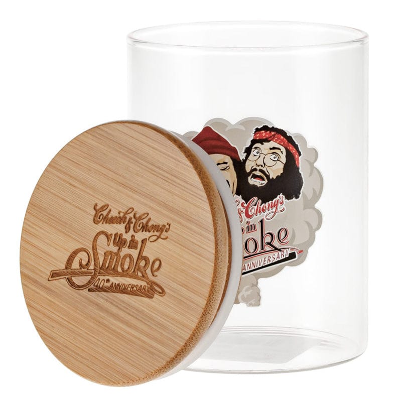 A three-quarter view of the C&C Heads Stash Jar - Mittel showing a clear glass cylinder with a leaning engraved bamboo lid and a colorful caricature label inside.