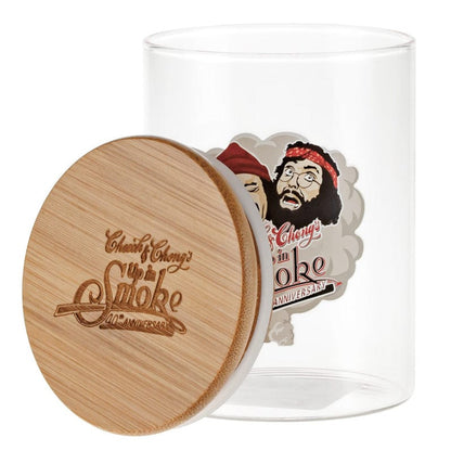 A three-quarter view of the C&C Heads Stash Jar - Mittel showing a clear glass cylinder with a leaning engraved bamboo lid and a colorful caricature label inside.