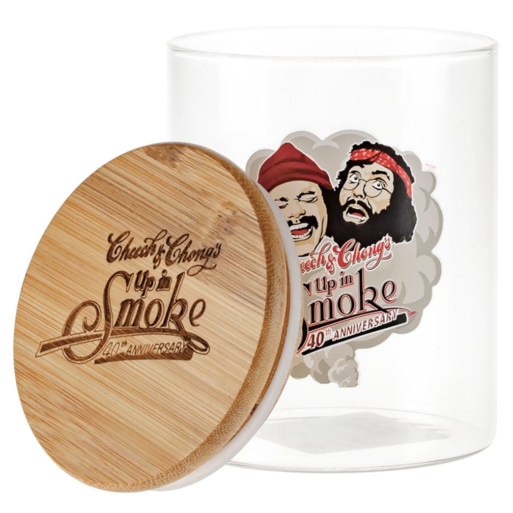 Angled view of the C&C Köpfe Stash Glas - Groß with a bamboo lid engraved "Up in Smoke" leaning against the clear jar showing a colorful Cheech & Chong label.