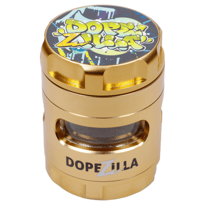 Angled close-up of the Dopezilla Space Pod 3-Stage 50 mm Grinder - Air showing its gold metal finish, graffiti-style logo top, and mesh viewing window.