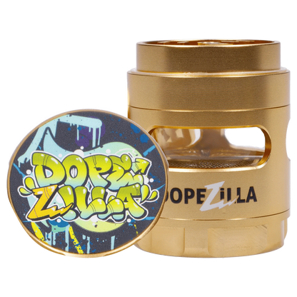 Close-up front angle of the gold Dopezilla Space Pod 3-Stage 50 mm Grinder - Air showing its stacked metal chambers and a colorful graffiti-style lid propped against it.