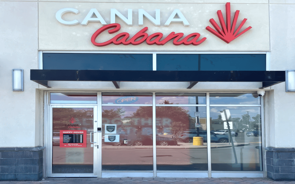 Canna Cabana storefront with glass doors and windows, red logo above, black awning, and store hours sign on the left door 