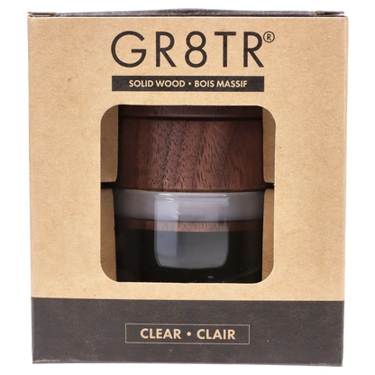 Close-up front view through the window of the RYOT - GR8TR Walnut Wood Top Grinder w/ Clear Glass Jar Body - 3 inch showing the dark walnut wood top and clear glass jar inside its cardboard packaging.