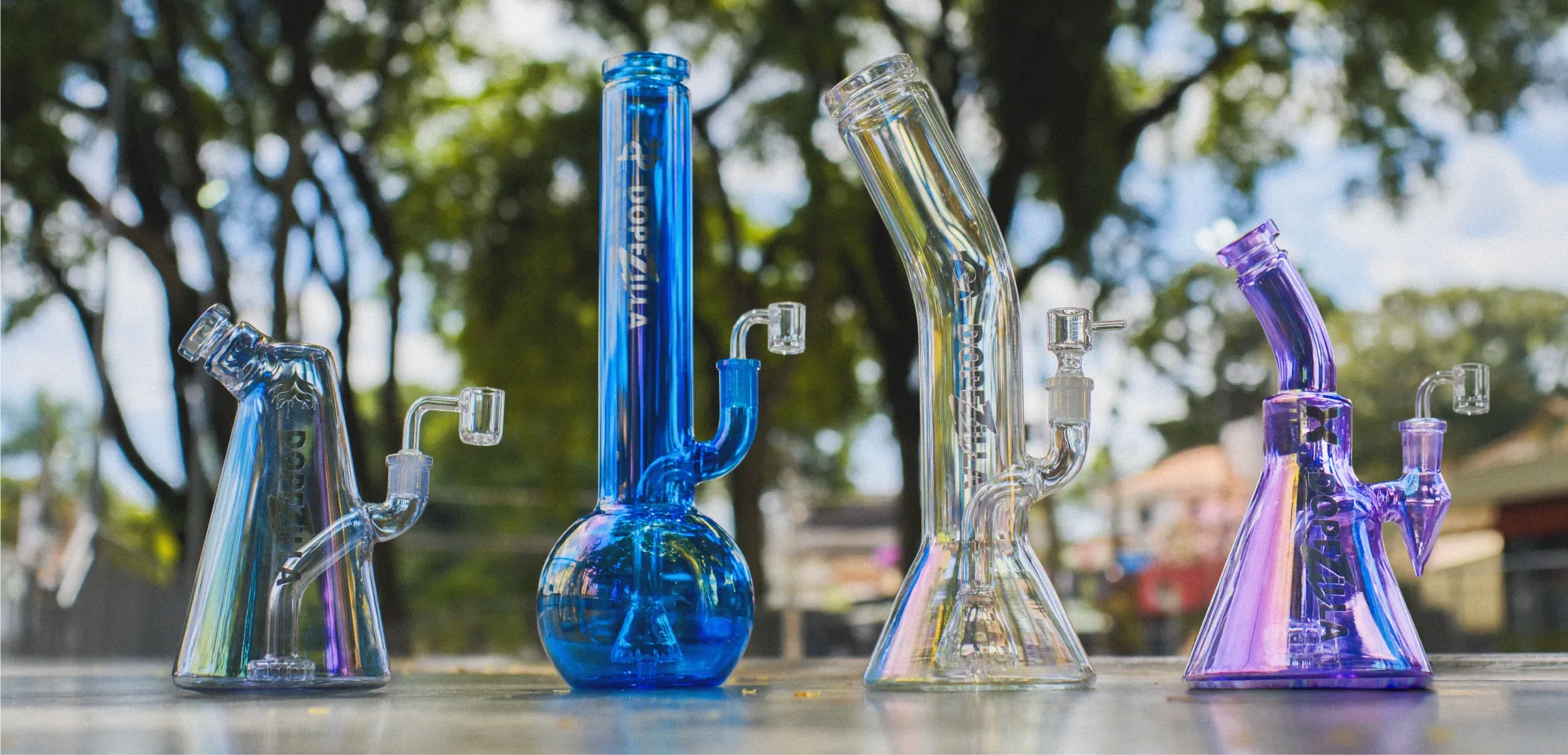 Four colorful Dopezilla glass rigs—iridescent, blue, clear, and purple—lined up on a table with blurred trees in background