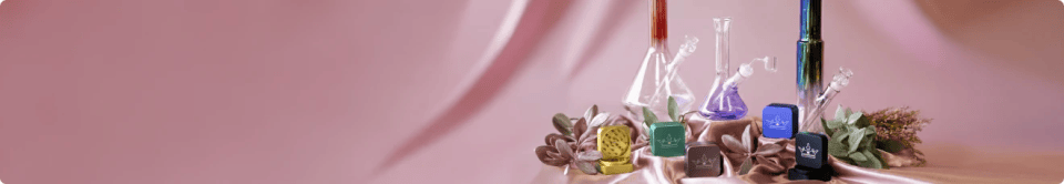 A row of colorful QOB grinders in front of glass bongs and succulents on a pink draped background.