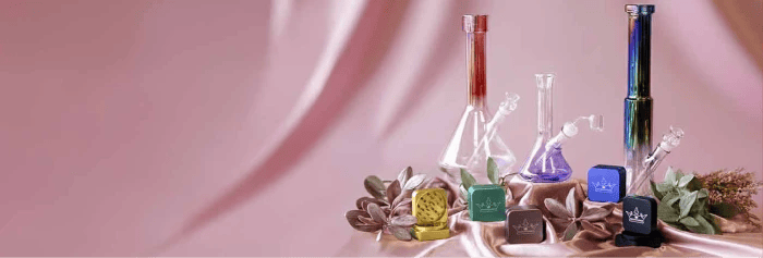 QOB bongs and grinders display: three glass bongs and five colorful QOB branded grinders arranged on silky pink fabric with decorative foliage.