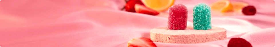 Two QŌB gummy pops—one pink, one teal—sugar-coated and standing on a round stone platform against a soft pink fabric backdrop