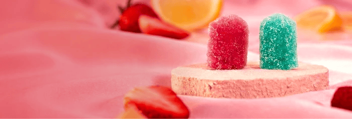 Two sugar-coated Qöb gummy chews (red and teal) standing on a pink pedestal with sliced strawberries and citrus on a pink backdrop