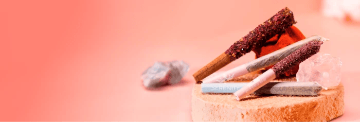 Three QOB pre-rolls—two coated in red kief and one plain—resting on a wood slice with crystals on a soft pink background