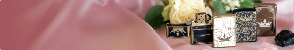 A row of decorated QOB Zippo-style lighters with crowns and floral designs arranged on pink fabric beside pale yellow roses